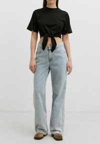 Mujer vistiendo un crop top negro de manga corta atado en la cintura, pantalones vaqueros de pierna ancha de color azul claro y zapatos marrones frente a un fondo blanco liso.