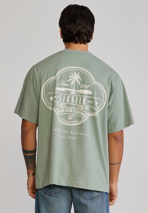 Férfi, aki zsályazöld pólót visel, fehér pálmafával és "SikSilk Beach Club & Lounge" logóval a hátán, kék farmerrel párosítva.