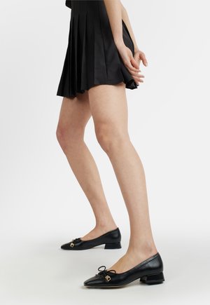 Donna che indossa scarpe basse nere in pelle con charm quadrati dorati, abbinate a una gonna corta nera a pieghe, in piedi su un pavimento bianco.