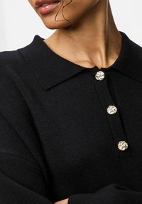 Melna džemperis ar klasisku apkakli, trim dekoratīviem sudraba tonī pogām un mīkstu, strukturētu audumu.