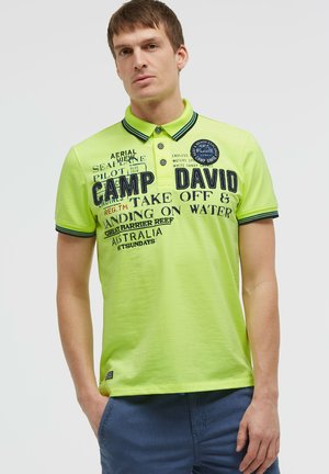 Man wearing a bright yellow-green polo shirt with "Camp David" and other text printed on it, and blue shorts with hands in pockets.