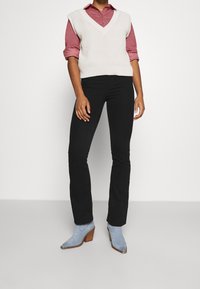 Licht beige gebreide vest over een rode overhemd met kraag, gecombineerd met zwarte uitlopende broeken en blauwe hakken. Glad textuur, gelaagde stijl.