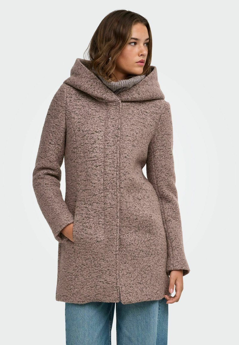 Femme portant un manteau à capuche mauve texturé à manches longues, les mains dans les poches, regardant sur le côté, avec un jean bleu en dessous.