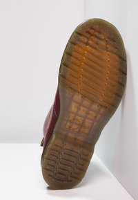 Semelle d'une botte rouge avec une bande de roulement en caoutchouc translucide et texturée, présentant des motifs antidérapants et des marquages de marque en relief.