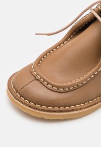 Pantofii din piele maro au un vârf semicircular, cusături decorative și o talpă de cauciuc ușoară. Include detalii de șireturi pe lateral.