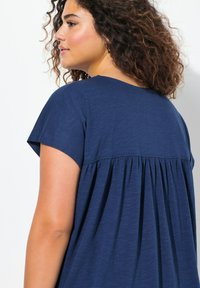 Námořnická modrá blůza s kulatým výstřihem, krátkými rukávy a nařaseným designem na zadní straně, vyrobená z lehkého, texturovaného materiálu.