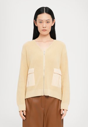 Light beige knit cardigan with a zip front, featuring two cream pockets and ribbed cuffs. Paired with brown trousers.
