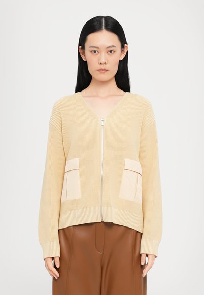 Hellbeige gestrickte Strickjacke mit Reißverschluss vorne, ausgestattet mit zwei cremefarbenen Taschen und gerippten Bündchen. Kombiniert mit braunen Hosen.