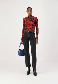 Rdeča in temnomodra cvetlična majica z dolgimi rokavi, združena z visokim pasom črnih kavbojk in gležnjaki. V desni roki je modra torbica.