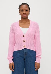 Gaiši rozā cropped kardigans ar V-veida kakla izgriezumu, atvērtu adījumu un trim lielām brūnām pogām. Iezīmē garas piedurknes un ribotas aproces.