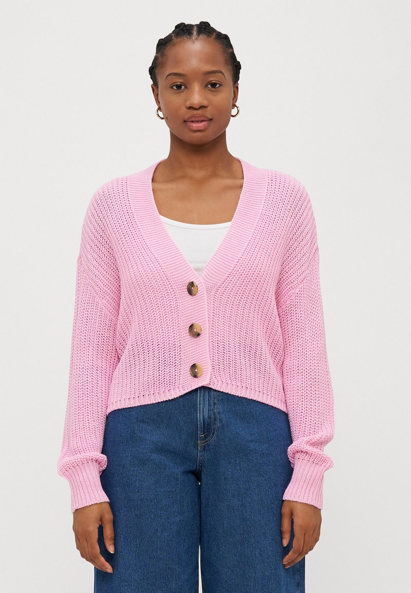 Gaiši rozā cropped kardigans ar V-veida kakla izgriezumu, atvērtu adījumu un trim lielām brūnām pogām. Iezīmē garas piedurknes un ribotas aproces.