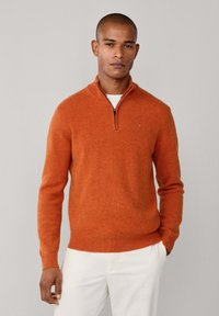 Pullover lavorato a maglia arancione con colletto a mezzo zip e polsini e orlo a coste. Il tessuto sembra morbido, con una finitura testurizzata. Indossato sopra una camicia bianca.