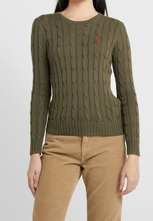 Personne portant un pull torsadé vert olive avec un logo rouge brodé et un pantalon en velours côtelé beige, debout devant un fond clair uni.