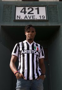 Jersey a rayas blancas y negras con el texto "ARISTON" y un parche de la bandera italiana, llevado con vaqueros azules, de pie frente a una puerta verde.