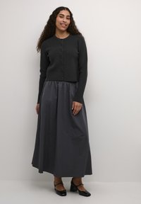 Cardigan tricotat de culoare gri închis, cu închidere cu nasturi, asortat cu o fustă maxi fluidă de culoare gri deschis. O pereche de pantofi cu toc bloc negri completează ținuta.