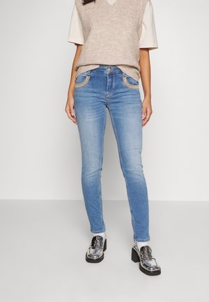 Jeans ajustados de color azul con un lavado claro, con detalles de costura en los bolsillos, combinados con un chaleco de punto beige y unos zapatos plateados brillantes.