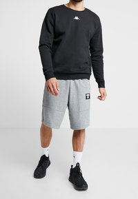 Man som bär en svart långärmad sweatshirt, grå knälånga shorts med Puma-märke, vita Puma-strumpor och svarta sportskor, stående på ett vitt golv.