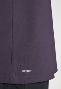 Fialová športová košeľa vyrobená z hladkej tkaniny, so zaobleným lemom a bielym logom "AEROREADY" blízko dolného lemu.