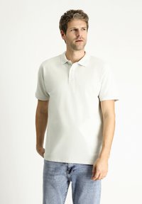 Polo shirt grigio chiaro con maniche corte, dotato di una chiusura a tre bottoni e un logo discreto sul petto. Abbinato a jeans in denim blu.