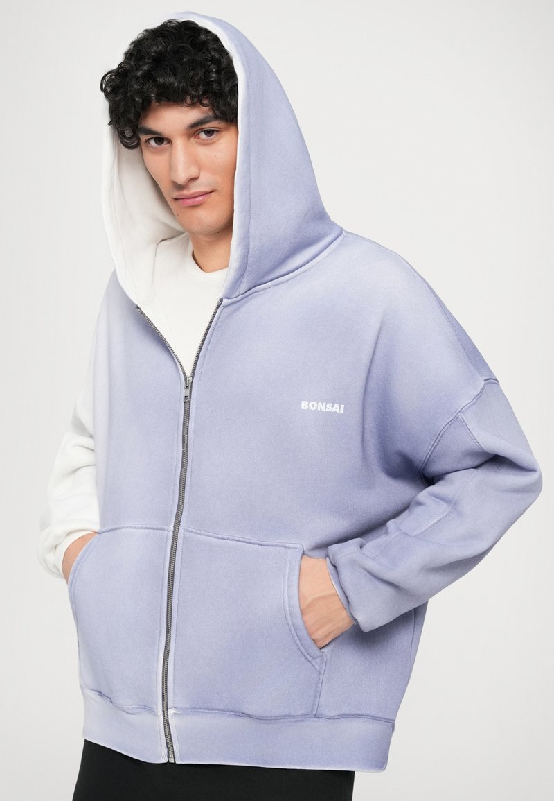 Lilla og hvid zip-up hoodie med frontlommer, blød tekstur og "BONSAI" logo. Har en stor hætte og afslappet pasform.