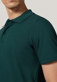 Ciemnozielona koszulka polo z klasycznym kołnierzykiem, krótkimi rękawami i teksturowanym materiałem. Zawiera detal z zapięciem na guziki.