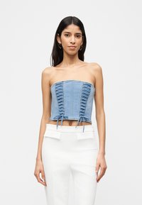 Γυναίκα που φοράει ένα στράπλες μπλε τζιν κορσέ τοπ με κορδόνια και λευκό παντελόνι ψηλής μέσης, σε απλό φόντο.
