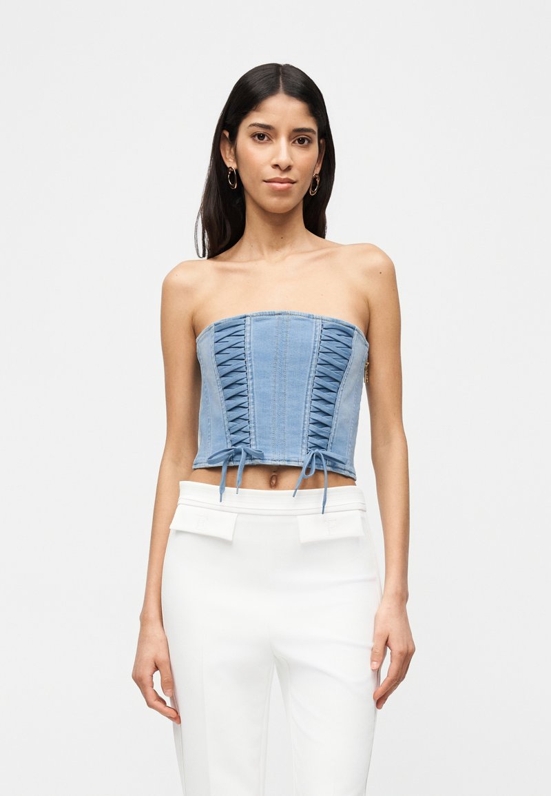 Γυναίκα που φοράει ένα στράπλες μπλε τζιν κορσέ τοπ με κορδόνια και λευκό παντελόνι ψηλής μέσης, σε απλό φόντο.