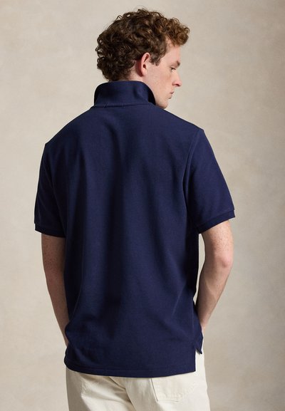 Muškarac sa kovrčavom kosom, nosi tamnoplavu polo majicu kratkih rukava i bijele hlače, okrenut leđima na bež pozadini.