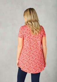 Túnica floral roja con mangas cortas, dobladillo redondeado y tejido suave, con pequeños patrones de flores rosas y blancas. Se lleva con leggins oscuros.