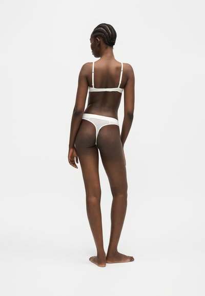 Femme aux cheveux tressés, debout pieds nus, portant un soutien-gorge blanc côtelé et un string, dos tourné sur un fond uni clair.