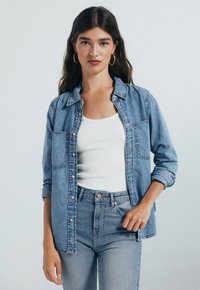 Denim overhemd met knoopsluiting in een ontspannen pasvorm, lichtblauwe kleur, twee borstzakken en opgerolde mouwen, gecombineerd met een witte tanktop en jeans.