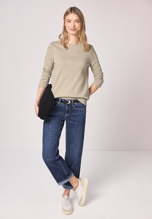 Femme debout, jambes croisées, portant un haut beige à manches longues, un jean bleu foncé retroussé, des baskets blanches, et tenant une pochette noire.