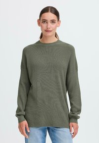 Olíva zöld, oversized pulóver, texturált kötött anyagból, hosszú ujjakkal és kerek nyakkivágással, világoskék farmernadrággal párosítva.