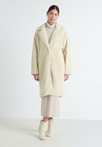 Krämsfärgad fuskpälsjacka med snibbkrage, långa ärmar och öppna fram. Bärs över en beige klänning och ankelstövlar.