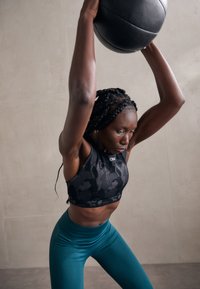 Črn športni crop top z visoko ovratnikom v vzorcu, v kombinaciji z visokimi teal leginami. Posameznik dviga črno medicinsko žogo.