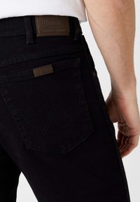 Jeans in denim nero con una toppa in pelle marrone che presenta la scritta "Wrangler", tasca posteriore contornata con cuciture a zigzag e design slim fit.