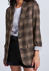 Brązowa kraciasta marynarka z pomarańczowymi i beżowymi akcentami, jednorzędowa, z dwoma guzikami, dopasowany fason, podwinięty mankiet z paskiem, noszona na tle graficznego t-shirtu.