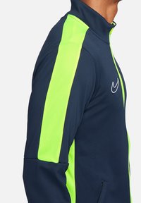Marineblauwe sportjack met opvallende neongroene zijstrepen, een hoge kraag en ritszakken, gemaakt van lichtgewicht, ademend materiaal.