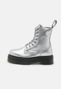 Stivaletto ankle in metallo argento con design a lacci, suola nera spessa e linguetta sul tallone. Texture liscia con forma della punta arrotondata.