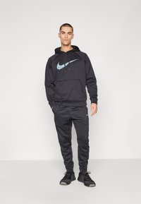 Juodas Nike džemperis su mėlynu ir baltu grafiniu logotipu; derinamas su juodais sportiniais kelnėmis, ant šlaunies turinčiomis mažą logotipą.