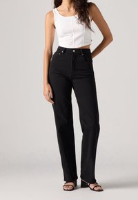 Femme portant un crop top blanc sans manches, un jean noir taille haute et des sandales noires à talons, debout devant un fond uni.