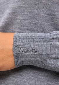 Szary mankiet swetra z dzianiny o prążkowanej fakturze i wyhaftowanym logo "Talk" kontrastującą nicią. Materiał wydaje się miękki i lekki.