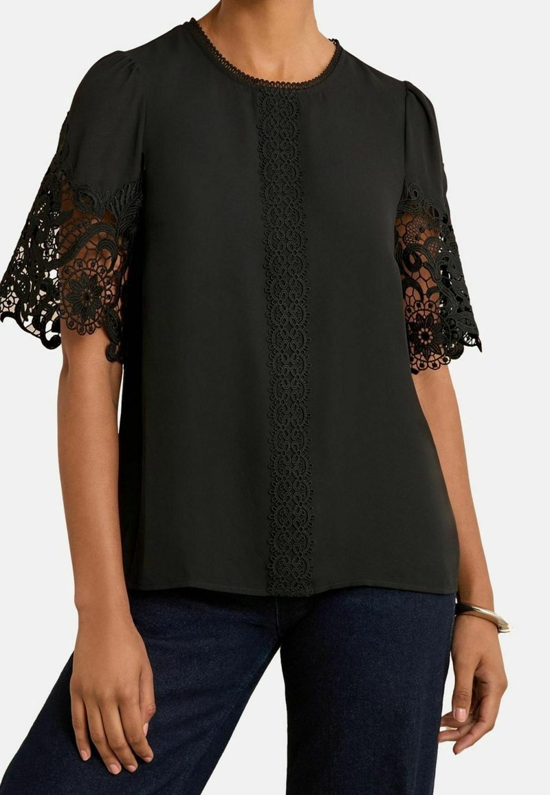 Blouse noire avec manches en dentelle complexe, col rond et un panneau de dentelle au centre. Tissu lisse avec une coupe décontractée.
