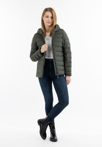 Jachetă puffer de culoare verde închis, cu glugă, design matlasat, cu fermoar pe față, purtată peste un top alb, asortată cu blugi bleumarin și botine negre până la gleznă.