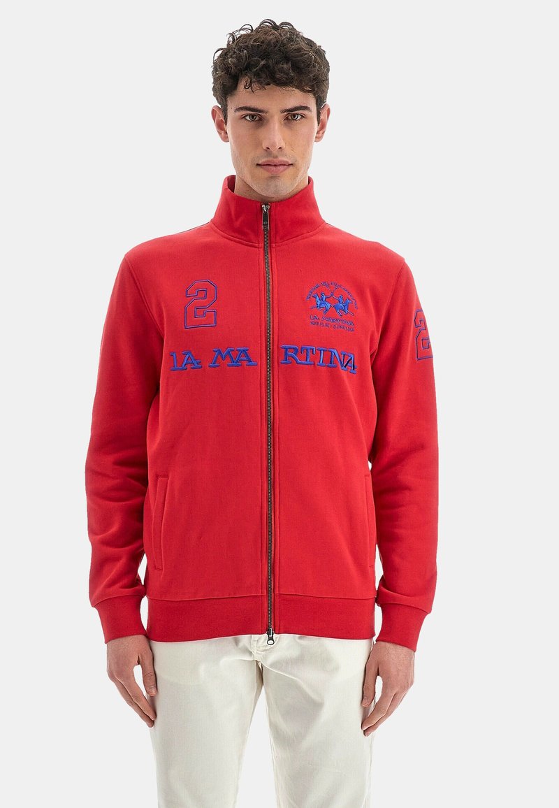 Giacca rossa con zip, maniche lunghe, con loghi e numeri ricamati blu. Realizzata in tessuto testurizzato con tasche laterali.