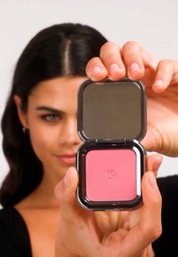 Colorete compacto en un estuche cuadrado negro reflectante. Contiene un polvo rosa suave, un logotipo en relieve y un espejo en la tapa.