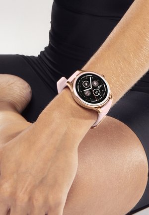 Smartwatch med et roseguldfarvet urkasse og rosa rem viser dato, skridt, puls og aktivitetsikoner på en persons håndled.