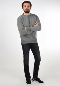 Pelēks džemperis ar apaļu kakla izgriezumu, ribotu aproci un oranžu logotipu uz krūtīm, komplektā ar melnām saplēstām džinsu biksēm un melnām sporta apaviem.