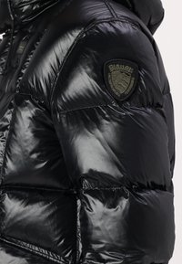 Jachetă neagră căptușită cu un finisaj lucios, având un badge cu logo pe umăr, buzunare cu fermoar și un design matlasat.