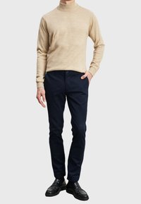 Maglione girocollo beige abbinato a pantaloni slim-fit blu navy e scarpe di pelle nere. Design semplici, texture morbida e colori in contrasto.